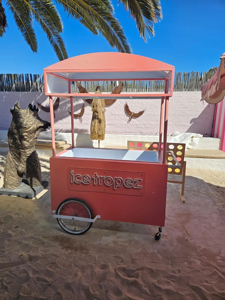 The original Ice Tropez cart on a beach under a palm tree, coral pink with a neon-lit sign