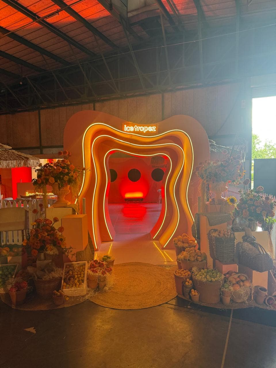 A large sculptural glowing tunnel entrance branded Ice Tropez, with produce displays and straw umbrellas at an indoor event